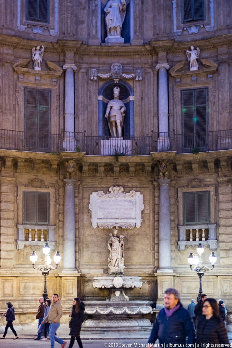 Pedestrians pass a decorated Palermo façade with balconies, pilasters, niche statues, and street lamps.