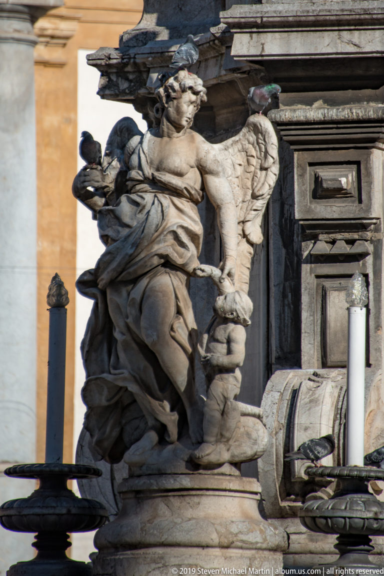 A crouching stone male figure supports a projecting element on an ornate façade.