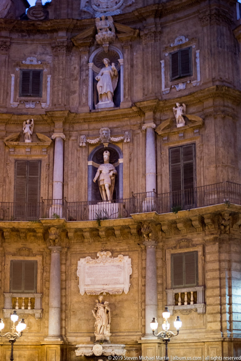 A pale statue stands in a façade niche above a carved panel framed by pilasters and volutes.