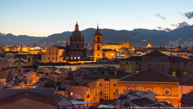 Illuminated church towers and a dome rise above dark roofs and Palermo’s night skyline.
