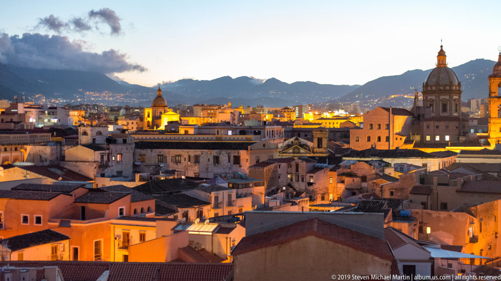Shadowed Palermo rooftops lead to illuminated domes and towers beneath a dark hillside.