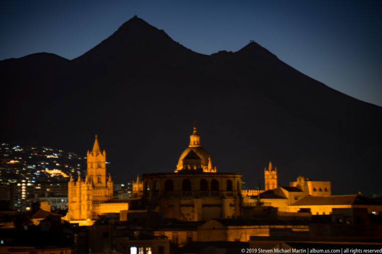 Warmly illuminated domes, towers, and a pointed spire rise above dark Palermo rooftops at night.