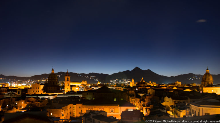 Church domes and towers silhouette against a blue dusk sky above Palermo’s emerging city lights.