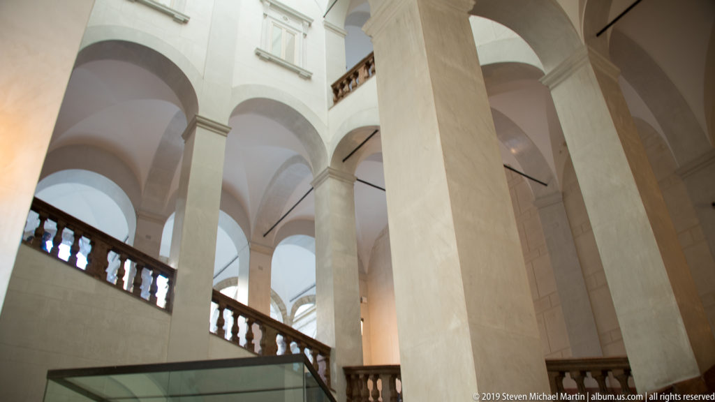 Stone staircase with dark balustrades rises among tall pale piers and rounded arches inside the Norman Palace.