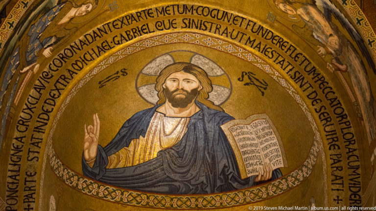 Gold mosaic of Christ Pantocrator blessing with his right hand and holding an open book within an inscribed arch.