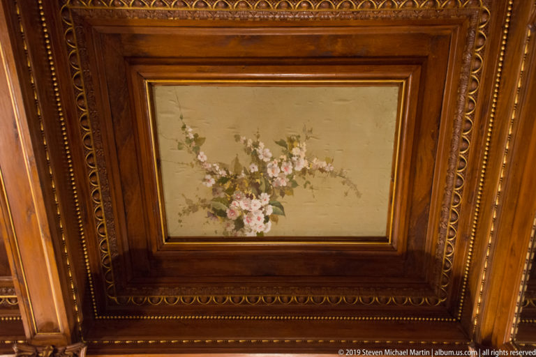 Pale floral painting with pink and white blossoms framed by richly molded polished wood paneling.