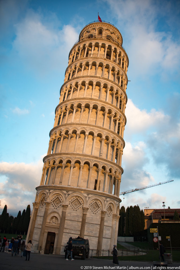 Full Leaning Tower of Pisa tilting across a blue sky, with stacked arcades glowing in evening light.