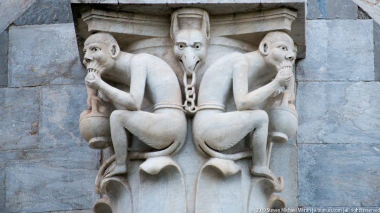 Close view of pale stone carving with two crouching figures flanking a long-eared central grotesque.
