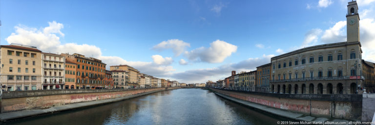 Arno River viewed from a bridge, lined by multistory palazzi and embankment walls through central Pisa.