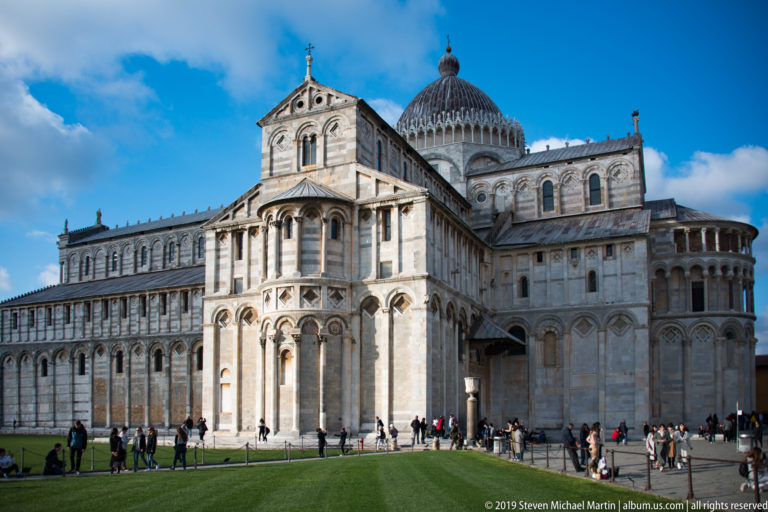 Pisa Cathedral seen across the lawn with its apse, transept, dome, marble arcades, and visitors along the base.