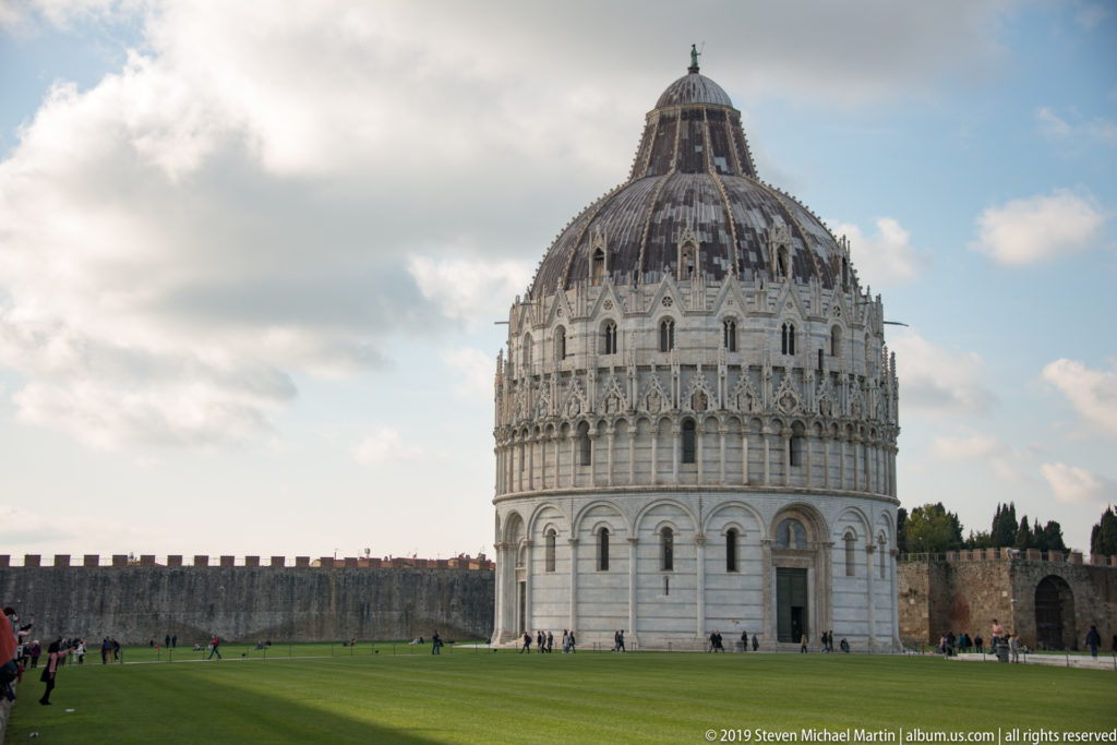 Pisa Baptistery centered on the green lawn, with its round marble walls, ornate upper tiers, dome, and city wall.