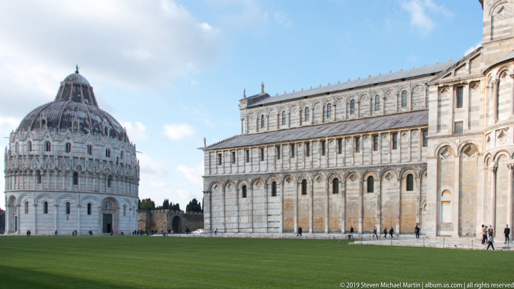 Pisa Baptistery and the marble side of Pisa Cathedral facing a broad green lawn under a blue, clouded sky.