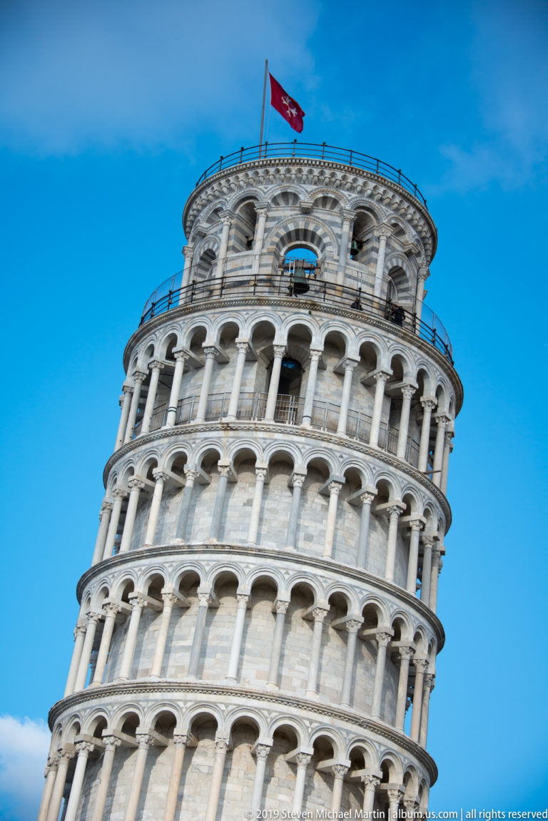 Close upward view of the Leaning Tower of Pisa showing tilted white marble arcades and the flag at its summit.