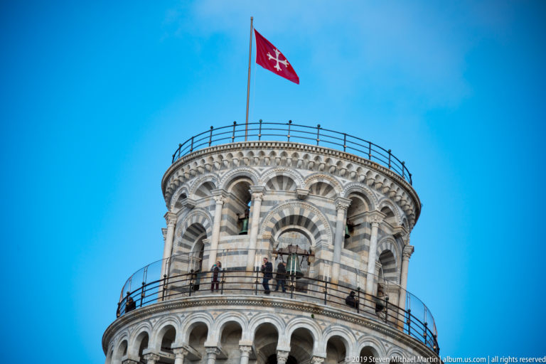 Upper Leaning Tower with open arcades, visitors on railed galleries, and a red Pisan flag flying above.