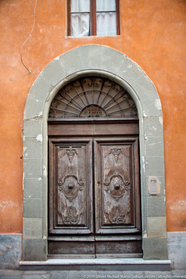 Dark carved wooden double door beneath a fanlight grille and gray stone arch in an orange stucco wall.