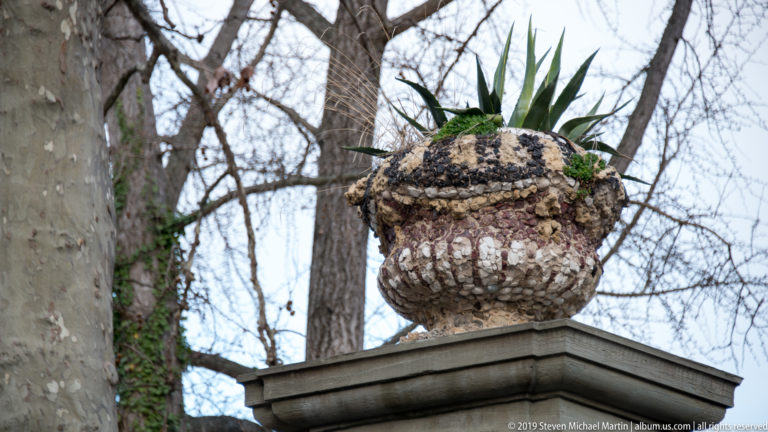 Weathered sculptural stone urn on a square pedestal planted with a spiky succulent among bare trees.
