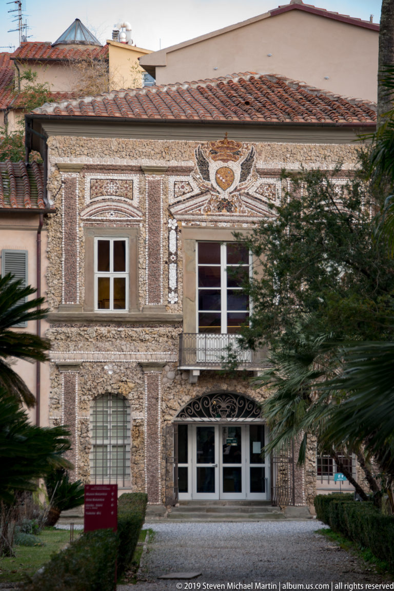 Symmetrical historic building with brick-and-stone patterned façade, central entrance, crest motif, palms, and small garden.