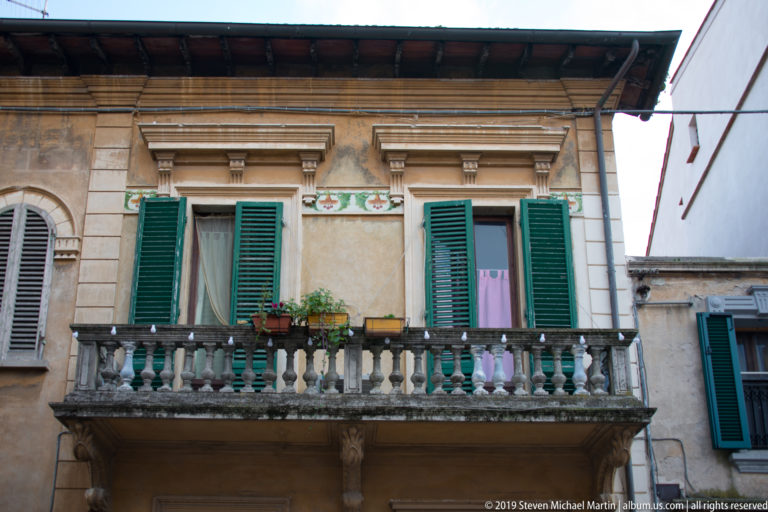 Stone balcony with balustrade, green-shuttered windows, potted plants, decorative panels, and laundry on a residential façade.