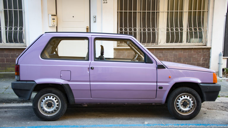 Lavender compact hatchback parked curbside before barred windows on a masonry building in Pisa.
