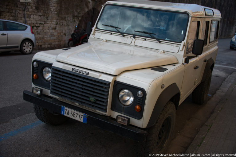 Cream Land Rover Defender with round headlights and black grille parked along a narrow street beside old masonry.