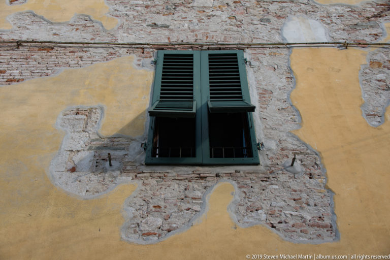 Green-shuttered window set in a weathered ochre wall with large patches of exposed brick and masonry.