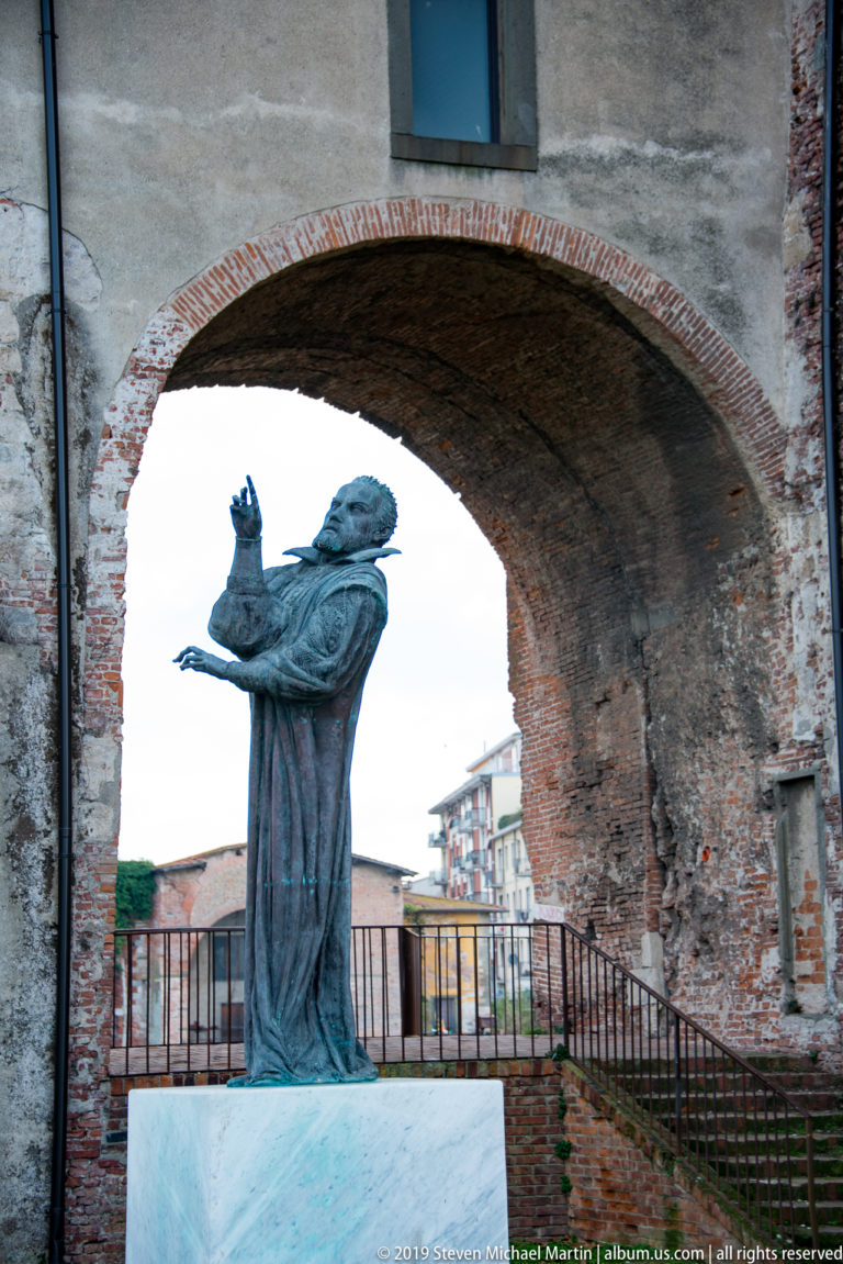 Bronze monument to Galileo Galilei standing on a pale pedestal beneath a brick arch at Pisa’s Cittadella.
