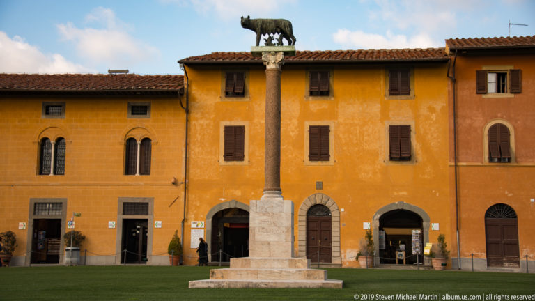 Stone column topped by the Capitoline Wolf and twins, standing before ochre historic buildings in Pisa.