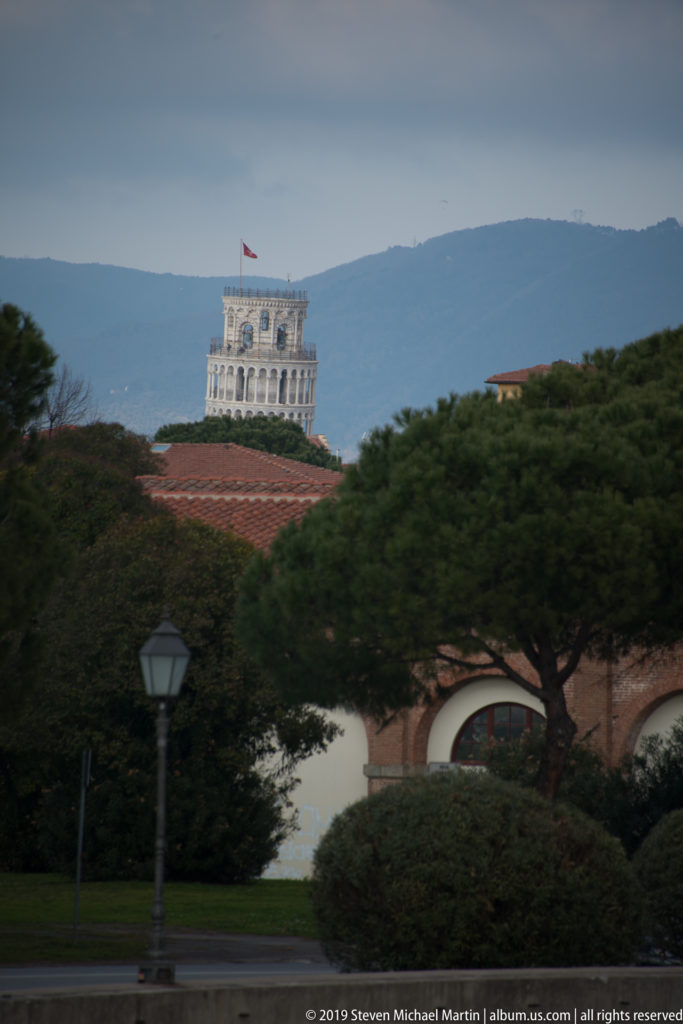 Upper white arcades of the Leaning Tower of Pisa visible above trees and red roofs against a distant mountain ridge.