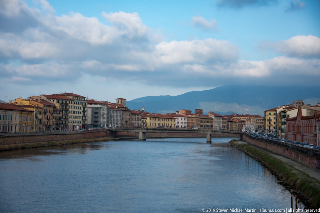 Arno River flowing between colorful riverfront buildings toward a low bridge, with mountains beneath clouds in the distance.