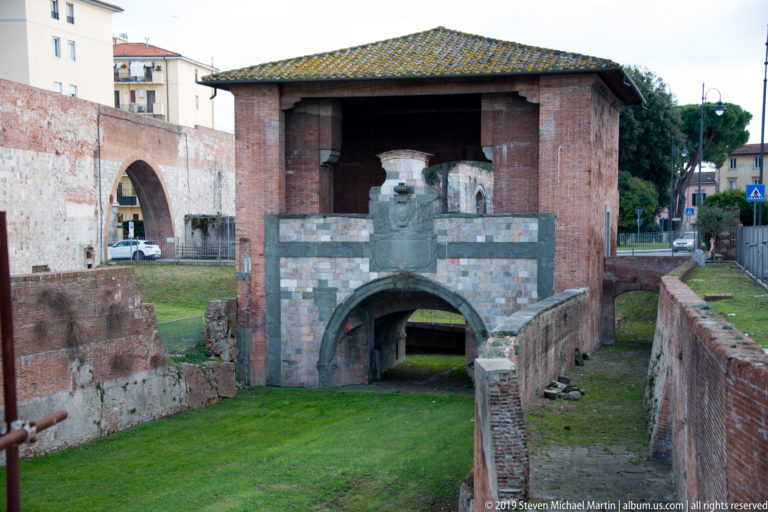 Weathered brick and pale-stone fortified gateway with arched passage, grassy interior, and adjoining historic walls.