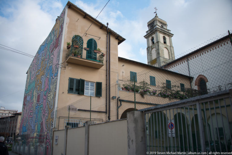Colorful mural on the side of a shuttered residential building with a pale bell tower rising behind it.