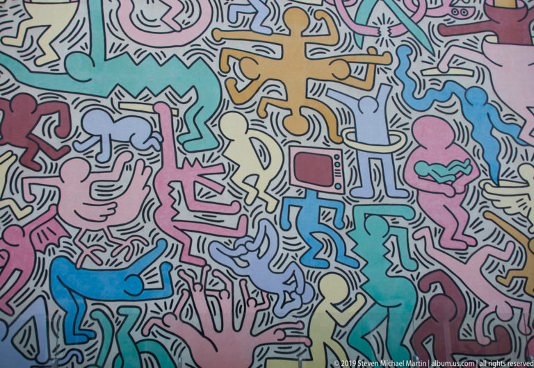 Wide mural section with a pink hand, blue animal-like form, and many interlocking human figures in pastel colors.