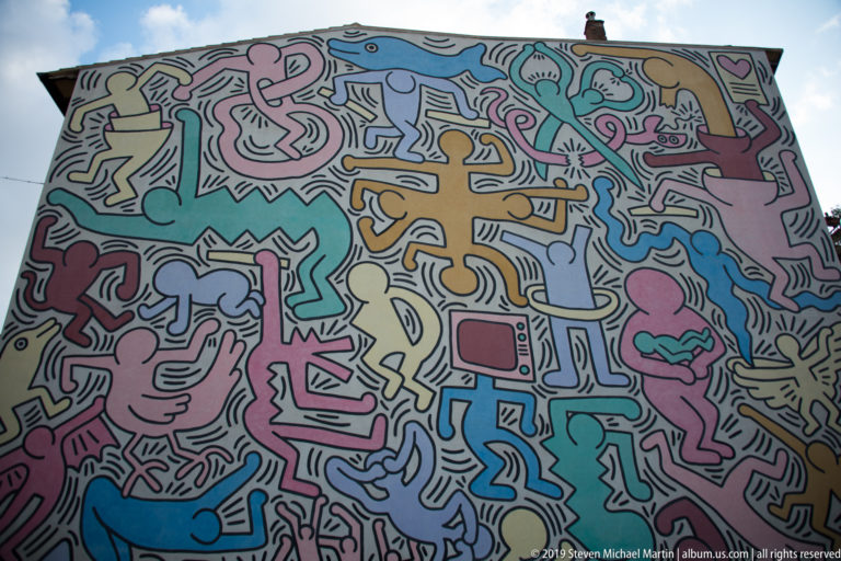 Broad view of a large multicolored mural filled with interlocking outlined figures, hands, animals, and symbols.