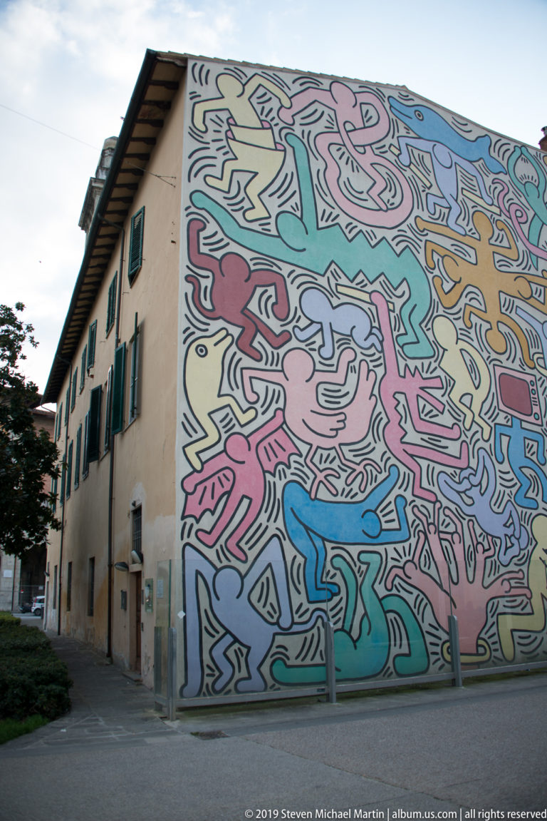 Oblique street view of a large colorful mural covering the end wall of a cream building with green shutters.