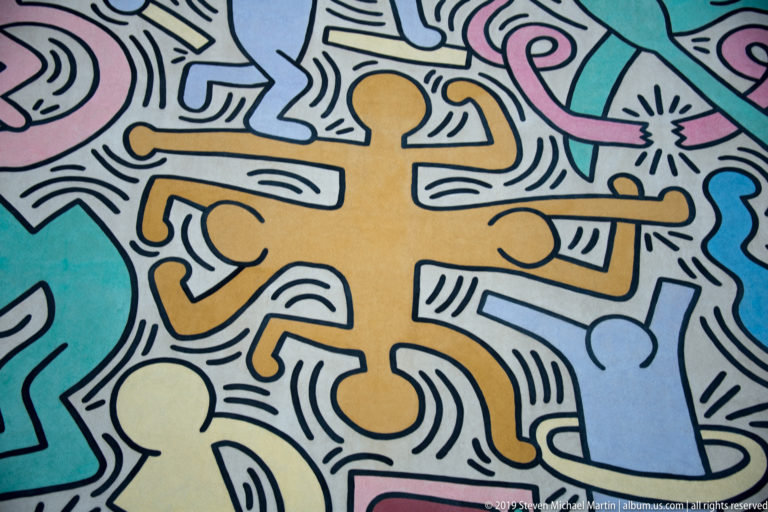 Orange running figure outlined in black amid yellow, blue, pink, and turquoise figures in a mural.