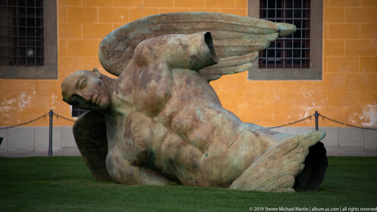 Large bronze winged human figure reclining on grass before an ochre wall in Pisa’s Piazza dei Miracoli.