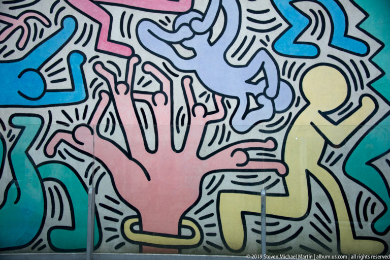 Large pink hand beside a yellow walking figure within a dense multicolored mural outlined in black.