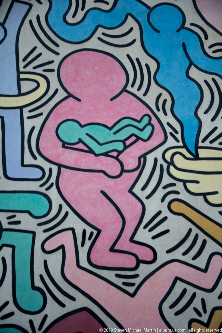 Pink crouching outlined figure holding a turquoise form amid blue, cream, and black linear mural elements.