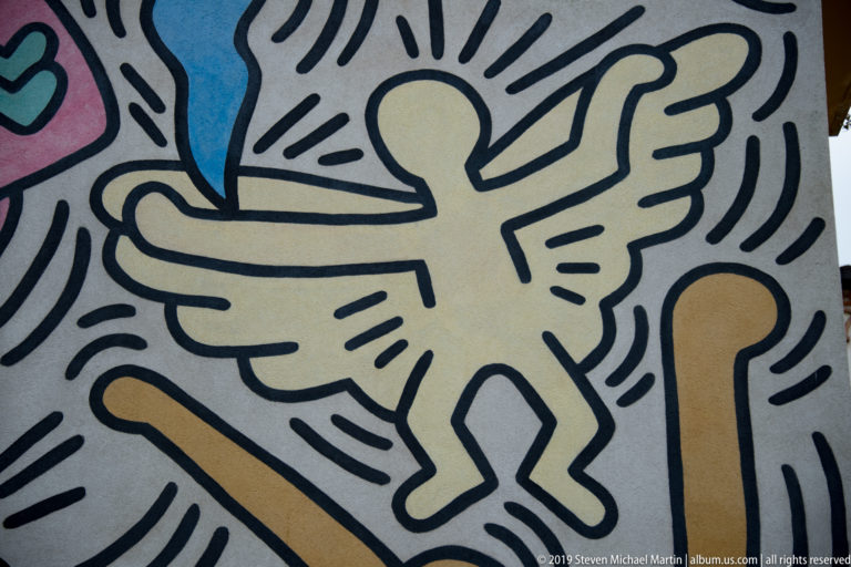 Close view of a pale yellow winged figure outlined in black within a colorful linear mural.