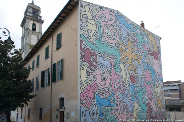 Large colorful mural of interlocking outlined figures covering the end wall of a shuttered building in Pisa.
