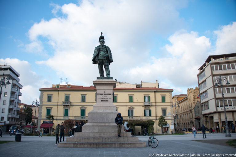 Bronze statue of Vittorio Emanuele II on a tall stone pedestal in an open Pisan square with pedestrians and bicycles.