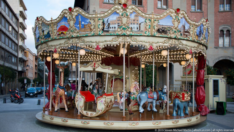 Decorative children's carousel with horses, carriage, mirrors, lights and red drapery standing among city buildings and scooters.