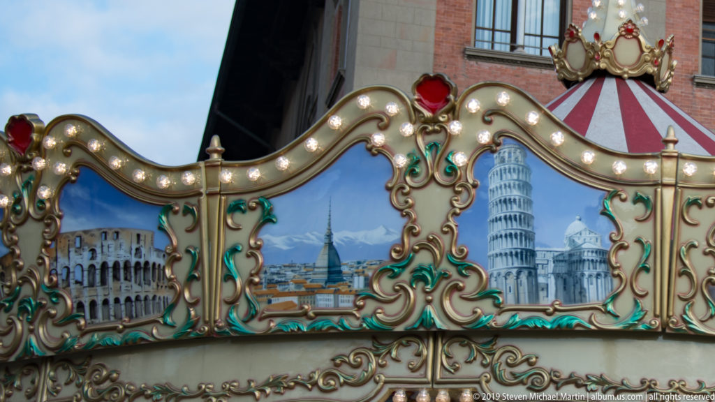 Ornate illuminated carousel panel painted with Pisa Cathedral and the Leaning Tower, framed by gilded scrollwork and a crest.