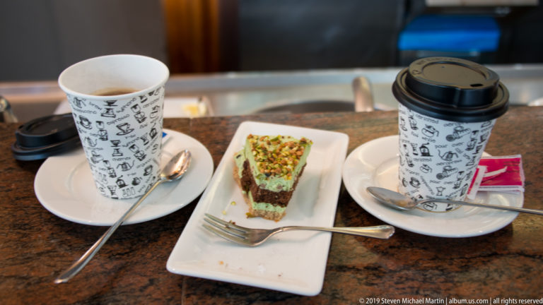 Two takeaway coffee cups and a small layered pastry topped with chopped green nuts on a café counter.