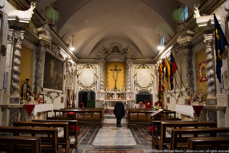 Wide view down a small decorated chapel toward a crucifix and altar, with benches, marble columns, paintings and flags.