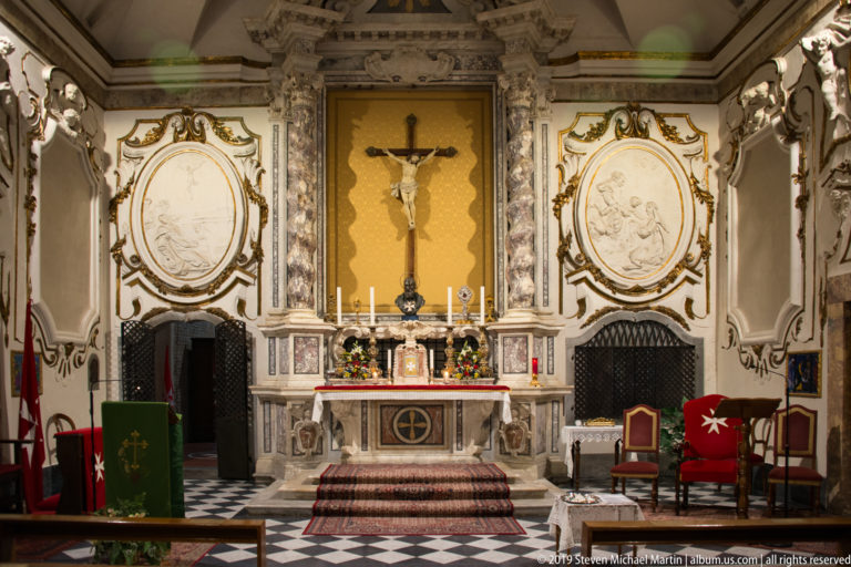 Ornate church altar with central crucifix, oval reliefs, candles, flowers, stucco decoration, and black-and-white floor.