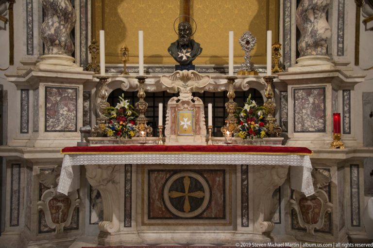 Marble chapel altar with flowers, tall candles, metal devotional objects and cross emblems on the tabernacle and frontal.