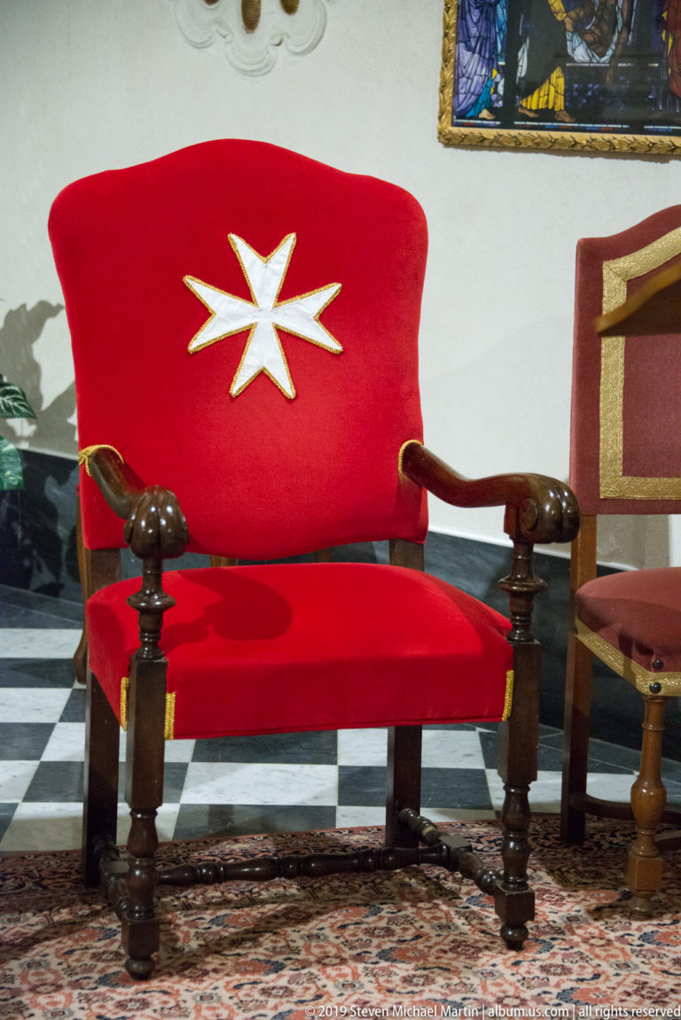 Red upholstered wooden armchair with a large white eight-pointed cross on the back, standing on a patterned rug.