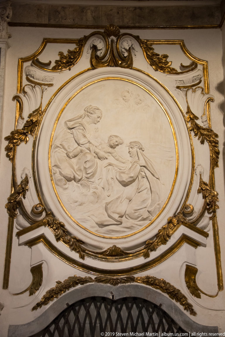 Oval pale relief of sacred figures surrounded by gilded scrollwork and painted architectural ornament above a dark grille.