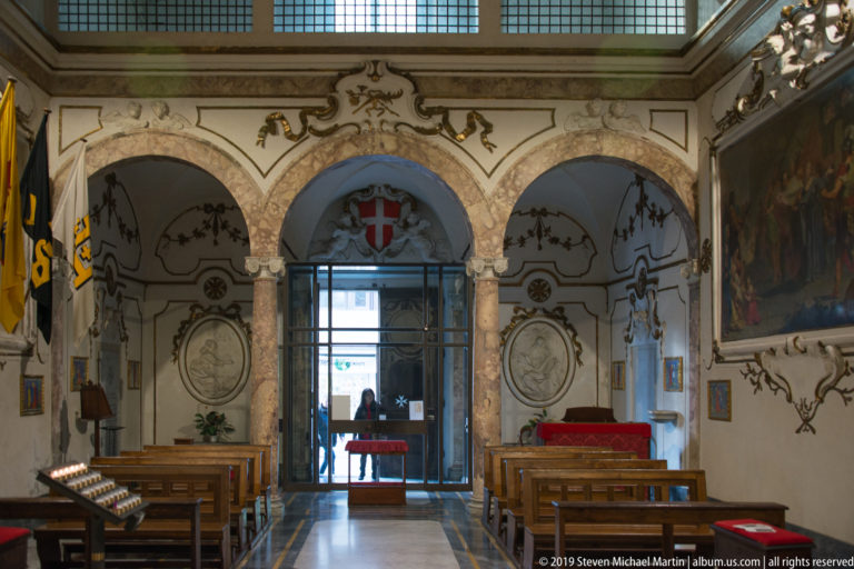 Chapel aisle leading to a bright doorway beneath a red shield with white eight-pointed cross, flanked by columns and benches.